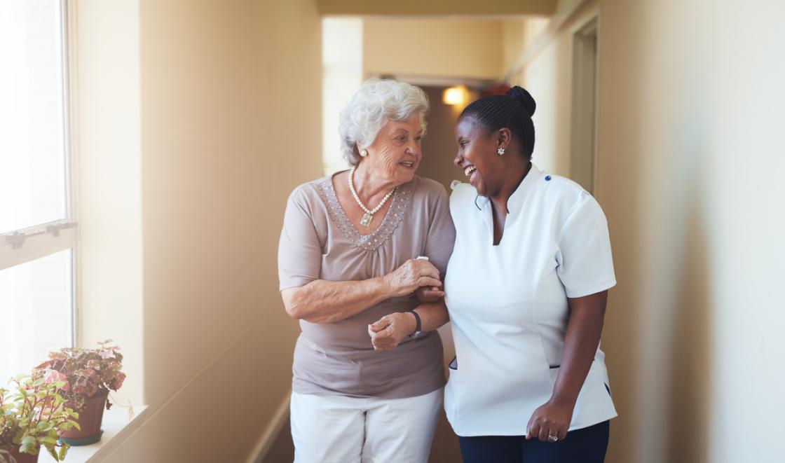 Older white woman walks down corridor support by black female care worker