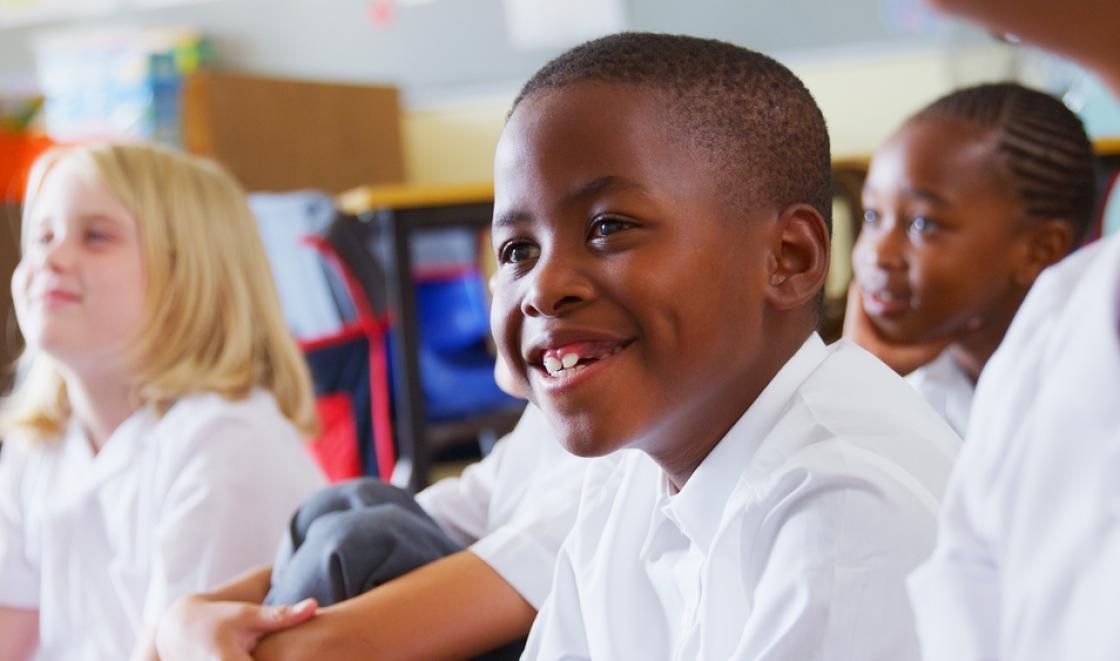 Black boy smiling in a classroom in the foreground with a white girl and black girl in the background